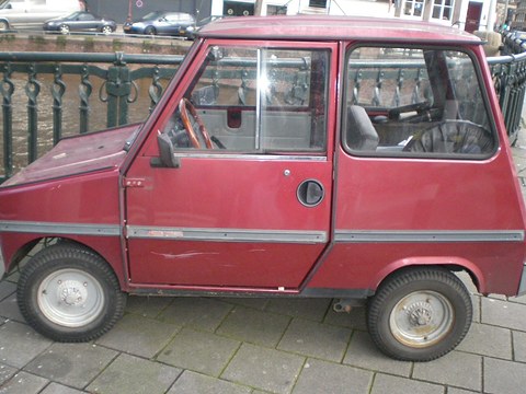 Side view of a boxy maroon two-seat microcar with large windows and small wheels beside a green iron railing. Opens a larger view