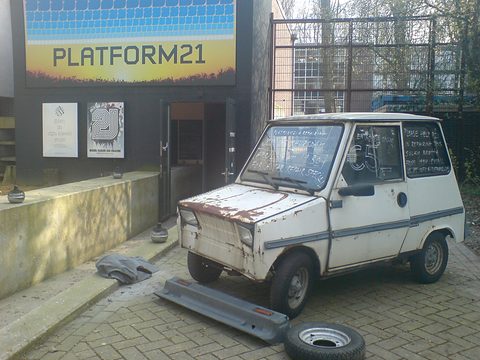 Rusting white boxy microcar with writing chalked on its windows, its bumper and a wheel lying on the paving beside it. Opens a larger view