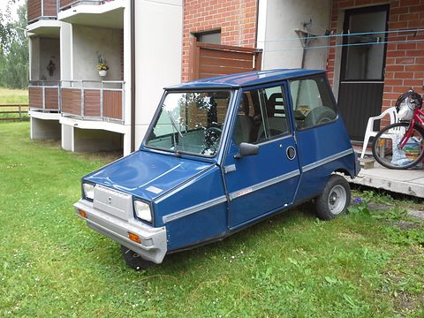 Blue boxy microcar with a grey front grille panel and chrome side strips, parked on grass in front of a block of flats.