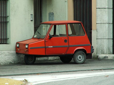 Side view of a small orange-red three-wheeled microcar with a sloping nose parked against a pale green wall. Opens a larger view