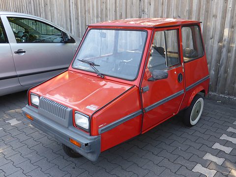 Red boxy microcar with a grey grille panel and bumper, square headlamps and white-wall tyres, parked on block paving. Opens a larger view