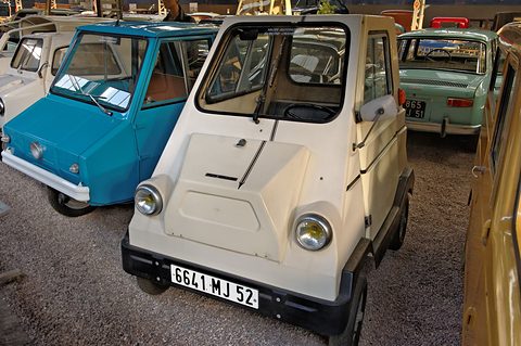 Cream four-wheeled boxy microcar with French plates and two round headlamps beside a turquoise Sulky, on gravel. Opens a larger view