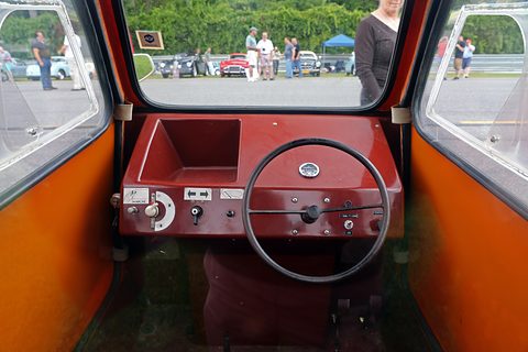 Spartan dark red dashboard with a thin black steering wheel, toggle switches and a choke labelled Marche, seen through the open doors. Opens a larger view