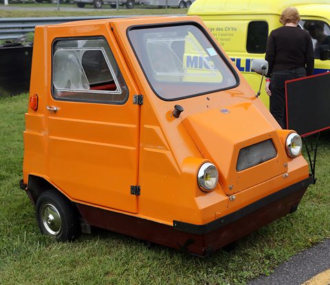 Orange three-wheeled Comtesse on grass at a concours, seen from the front three-quarter. Opens a larger view