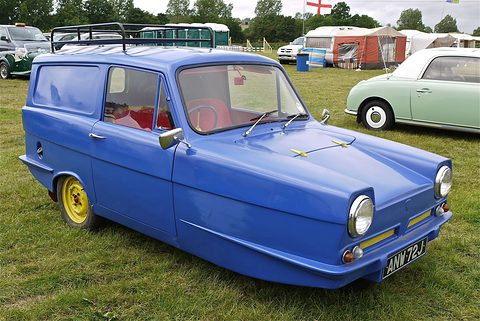 Bright blue three-wheeled van with yellow wheels and a roof rack on grass, caravans behind. Opens a larger view