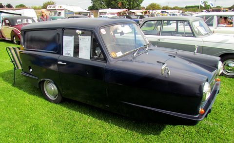 Navy three-wheeled van with notices in its window, parked on grass among classic cars. Opens a larger view