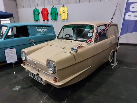 Cream three-wheeled van with bunting and a Union flag on a show stand, a blue van beside it. Opens a larger view