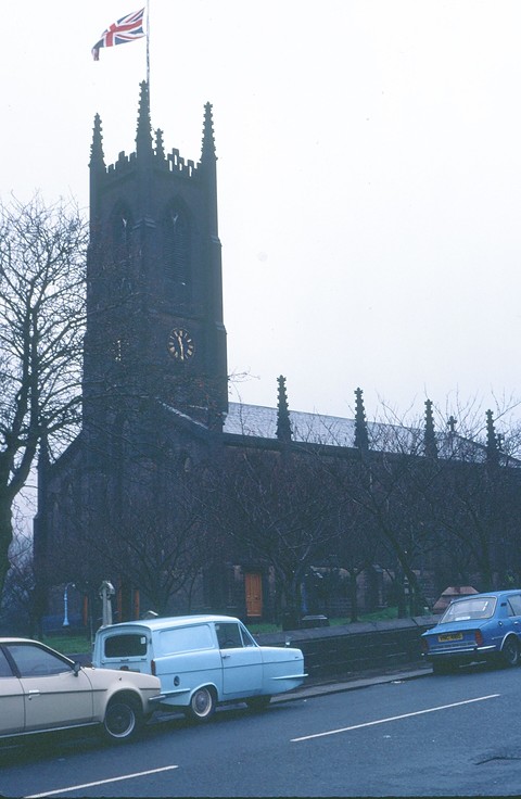 Dark church tower above a street of parked cars, a small pale blue van among them. Opens a larger view