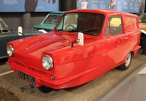 Red three-wheeled van with round headlamps and a smooth nose, on a museum floor beside other cars.