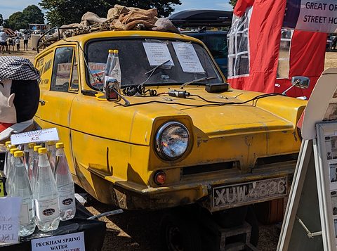 Yellow van head-on surrounded by props: bottled water, a cap and a figure in a flat cap. Opens a larger view