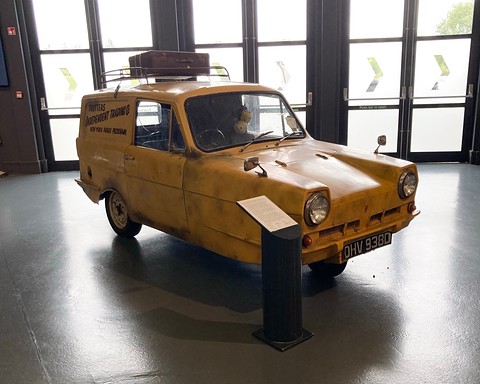 Yellow three-wheeled van on a grey floor before tall windows, a small display post beside it. Opens a larger view