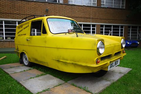 Grimy yellow three-wheeled van on paving slabs and grass in front of a brick building. Opens a larger view