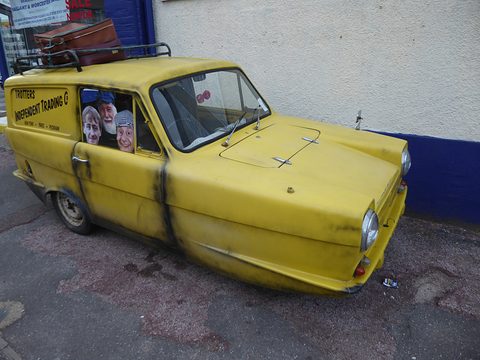 Grimy yellow van with two masked dummies at the side window, parked by a white wall. Opens a larger view