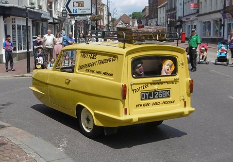 Yellow van with masked dummies in its windows on a high street, festival-goers behind. Opens a larger view