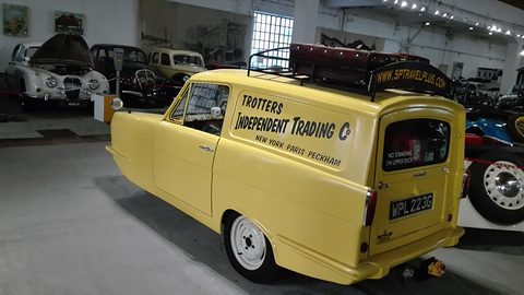 Yellow signwritten van from the rear quarter in a museum garage among classic cars. Opens a larger view
