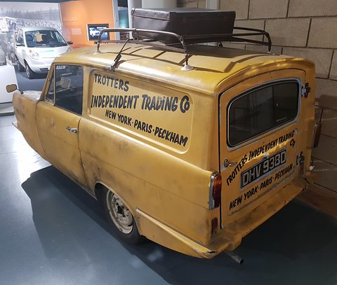 Yellow van from the rear quarter, lettered doors and a suitcase on the roof rack, in a museum hall. Opens a larger view