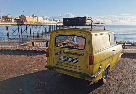 Grimy yellow van from the rear quarter on a seafront, a pier and calm sea behind. Opens a larger view