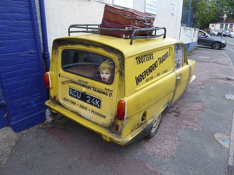 Yellow van from the rear quarter beside a blue wall, a masked dummy in the rear window. Opens a larger view