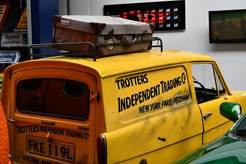 Back of a yellow van lettered Trotters Independent Trading Co, New York, Paris, Peckham, a suitcase on the roof. Opens a larger view