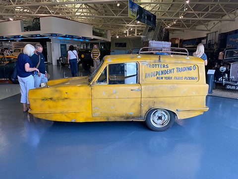 Weathered yellow van side-on on a blue-grey floor, visitors looking at exhibits behind. Opens a larger view