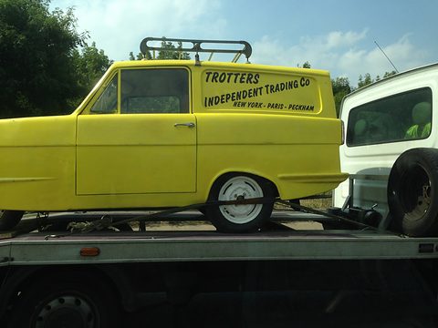 Yellow signwritten van side-on on the bed of a recovery lorry, trees behind. Opens a larger view