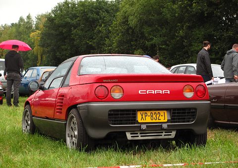 Rear of a red small coupé with CARA lettering and a yellow Dutch plate on grass, people standing around. Opens a larger view