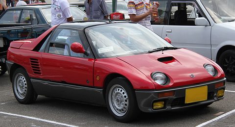 Red and grey small coupé with yellow fog lamps in the bumper and a yellow plate in a car park.