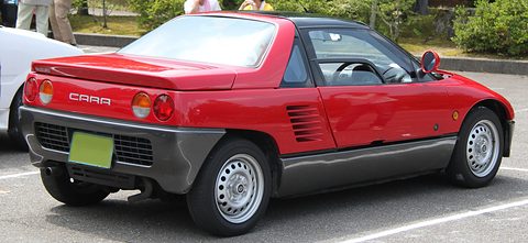 Rear of a red and grey small coupé with round tail lamps and CARA script, parked in a car park. Opens a larger view