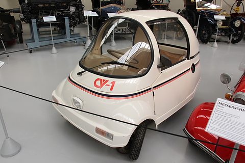 Tiny white single-seat car with a large glazed canopy and red CV-1 lettering, behind a rope beside a red bubble car. Opens a larger view