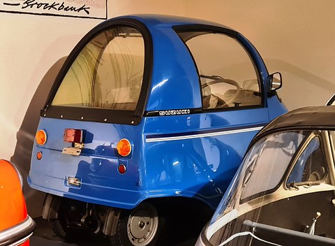 Rear quarter of a tiny blue single-seat car with a curved canopy, small round tail lamps and SUZUKI lettering, between other cars.