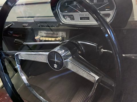 Black steering wheel with a chrome horn bar and Suzuki S badge in front of a strip speedometer reading to 100 km/h. Opens a larger view