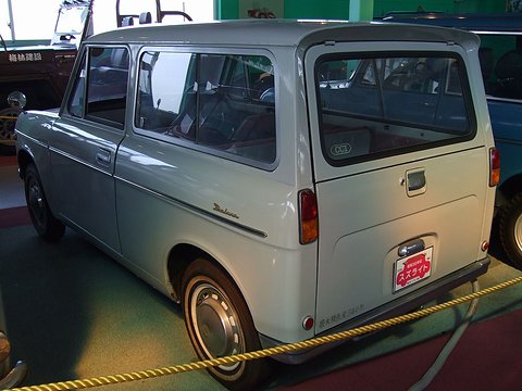 Rear three-quarter of a pale grey small van with a lift-up rear door and Deluxe script, behind a yellow rope. Opens a larger view