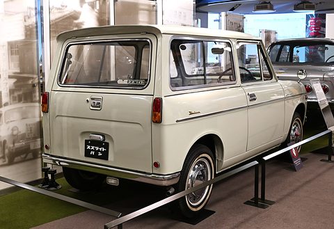 Rear three-quarter of a white small van with a large rear window, round tail lamps and a black plate behind a rail. Opens a larger view