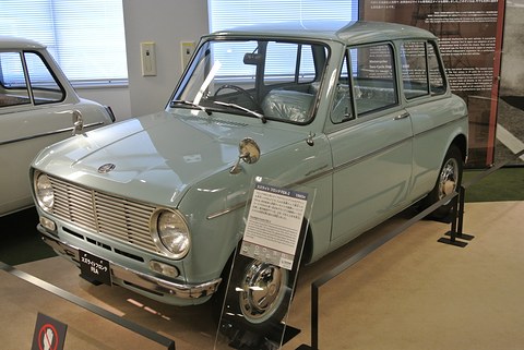Pale blue small saloon with a horizontal-bar grille, round headlamps and fender mirrors, a label stand by the front wheel. Opens a larger view