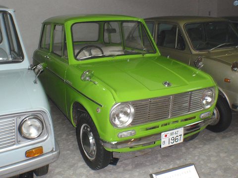 Bright green small saloon with a chrome grille, round headlamps and a 1967 plate between two other parked cars. Opens a larger view