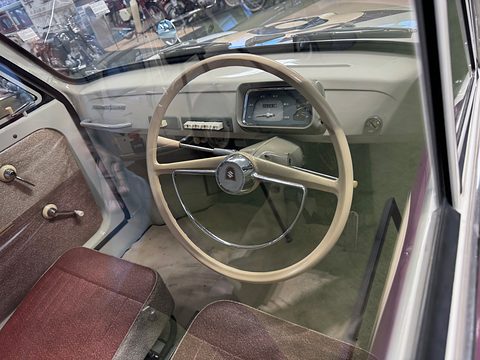 Cream two-spoke steering wheel with a chrome horn ring in front of a cream dashboard with a rectangular speedometer. Opens a larger view