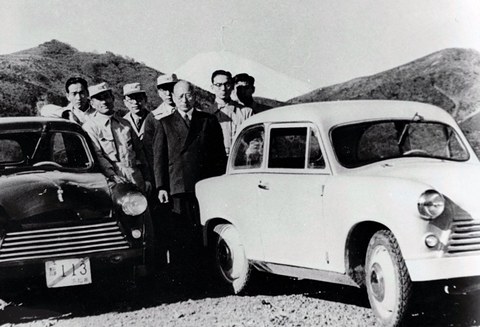 Black-and-white photograph of a group of men in coats standing between a dark and a light small saloon, a snow-capped peak behind.