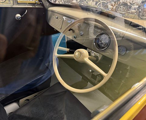 Cream painted dashboard with a single round dial and a large cream two-spoke steering wheel above black seats. Opens a larger view