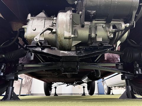 Underside view of a small engine and gearbox mounted crosswise above a leaf spring, with drive-shaft gaiters to each side.