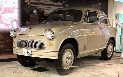 Beige small two-door car with a split vertical-bar grille in front of a painted street backdrop. Opens a larger view