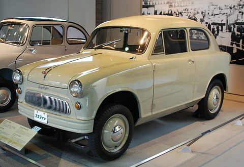 Cream two-door small car with a barred grille and round headlamps on a museum floor beside a label board. Opens a larger view