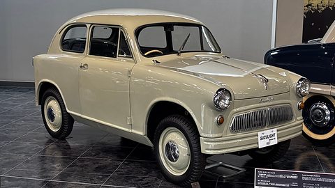 Beige two-door small car with white-wall tyres and a SUZULIGHT plate on a dark tiled floor. Opens a larger view