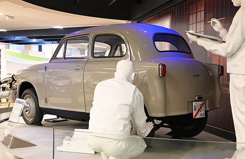 Rear three-quarter of a beige small saloon with a white kneeling figure at the rear wheel and a standing figure with a clipboard. Opens a larger view