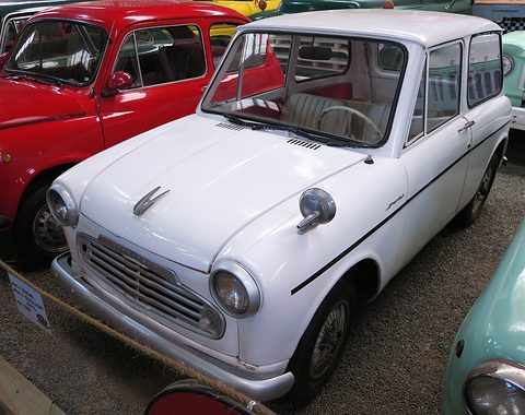 White small two-door van with a chrome grille, a bonnet mascot and a fender mirror, parked on gravel between other old cars. Opens a larger view