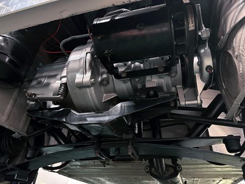 Close view from below of a small engine with a black dynamo, a gearbox casing and a transverse leaf spring. Opens a larger view