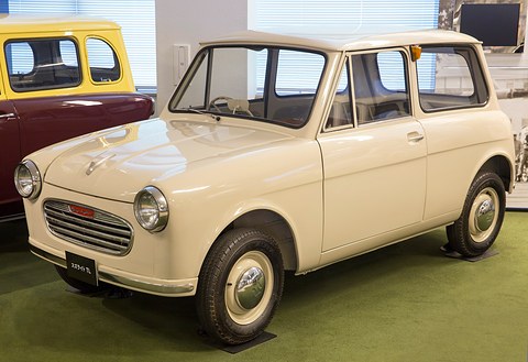 Cream boxy small van with round headlamps, a horizontal-bar grille and wheel covers, on green carpet.