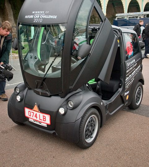 Small black three-seat city car with its whole front canopy raised, on a seafront road.