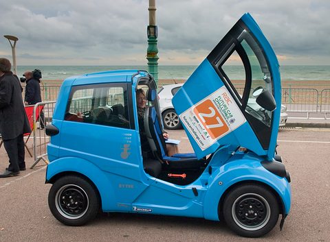 Blue small electric car with its forward-hinged door lifted, the driver’s seat visible.