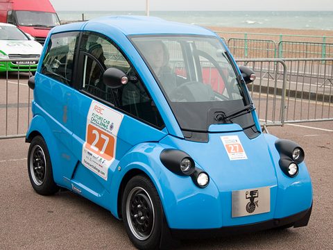 Bright blue small electric car with competition number 27 on its door, parked by railings.