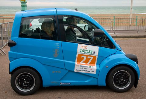 Blue small electric car side-on with an RAC Future Car Challenge sticker, sea behind. Opens a larger view